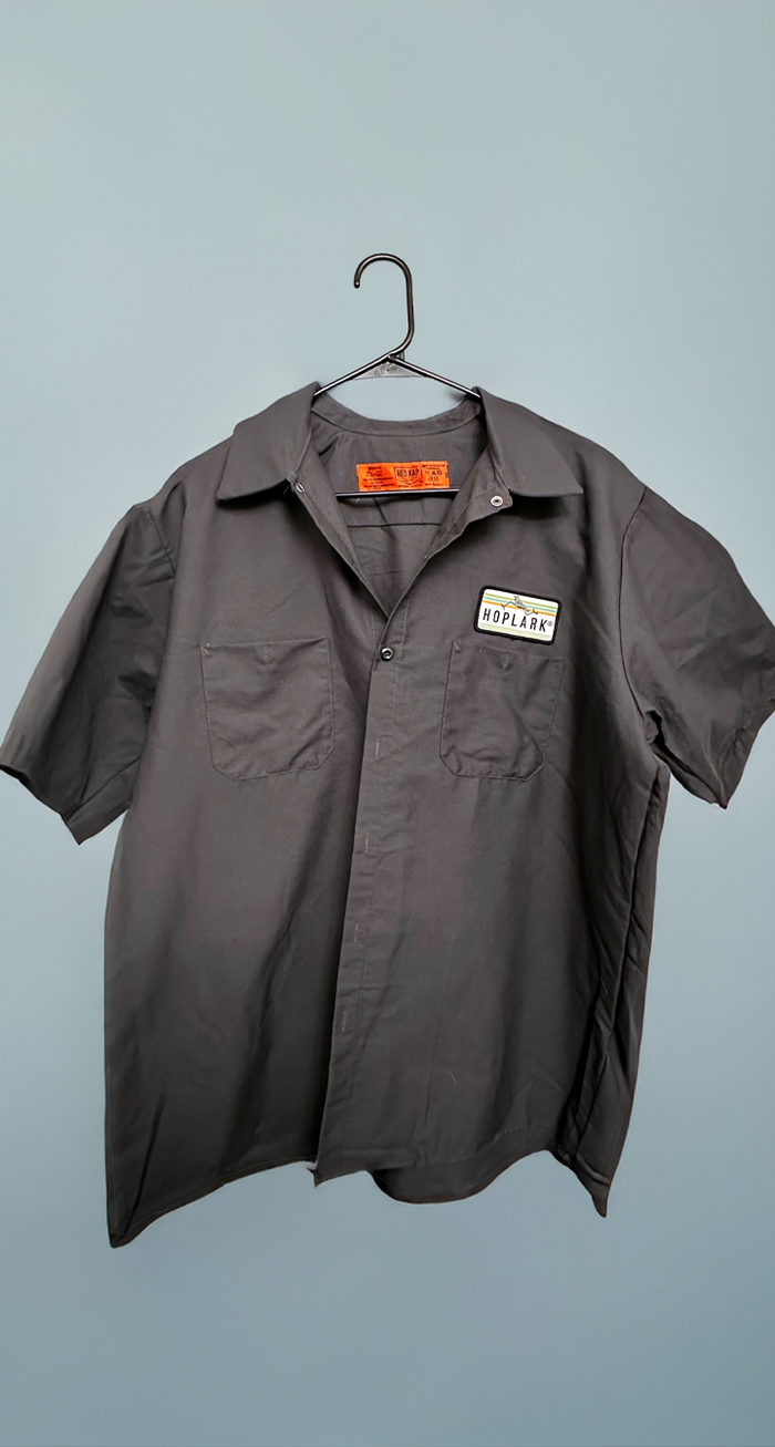 Gray short-sleeve shirt with hoplark patch, on a hanger against a light blue background